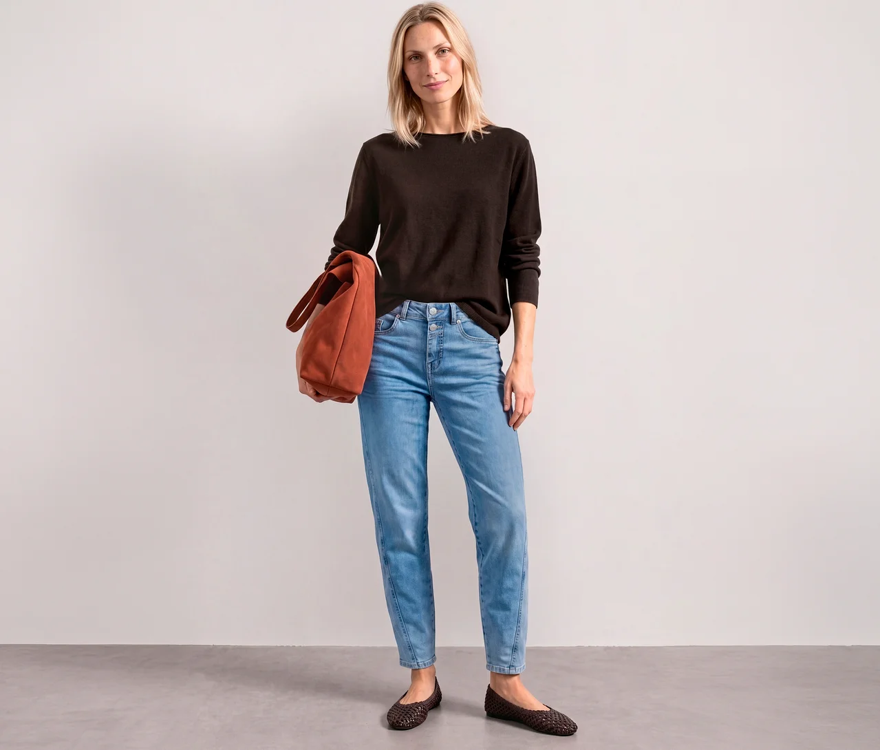 Une femme porte un pull en maille Street One marron, un jean bleu clair et tient un sac cabas orange.