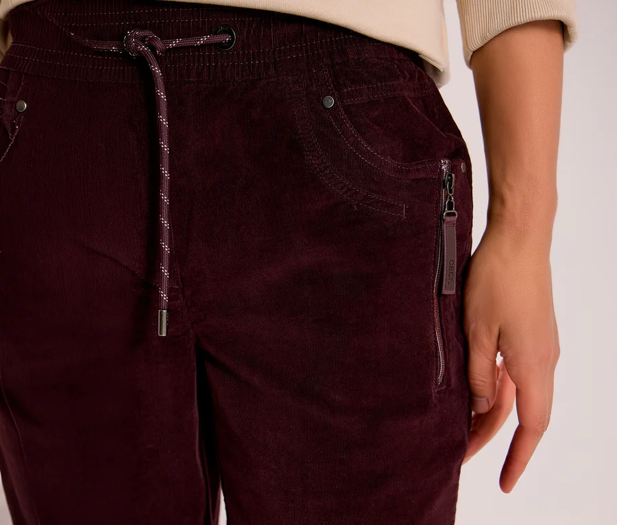 Gros plan d'une personne portant un pantalon en velours côtelé CECIL bordeaux, avec une taille élastique, un cordon de serrage moucheté et une poche latérale zippée avec une étiquette CECIL.