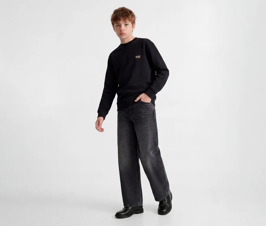 Jeune garçon debout, portant un sweat-shirt noir avec un petit logo doré, un jean gris foncé à jambes larges et des chaussures noires. Une main dans sa poche.