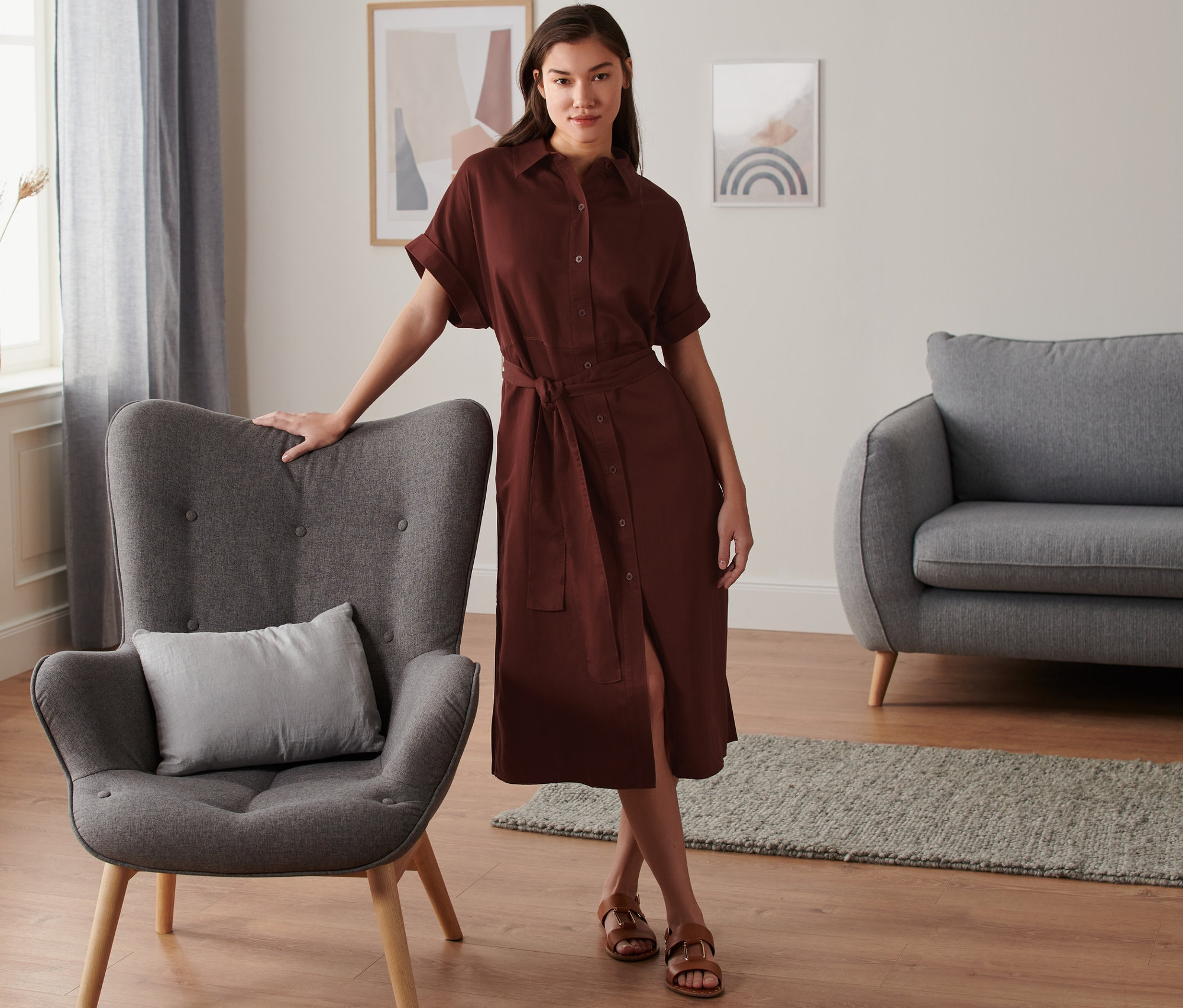 Femme posant en robe chemise marron en lin à côté d'un fauteuil gris.