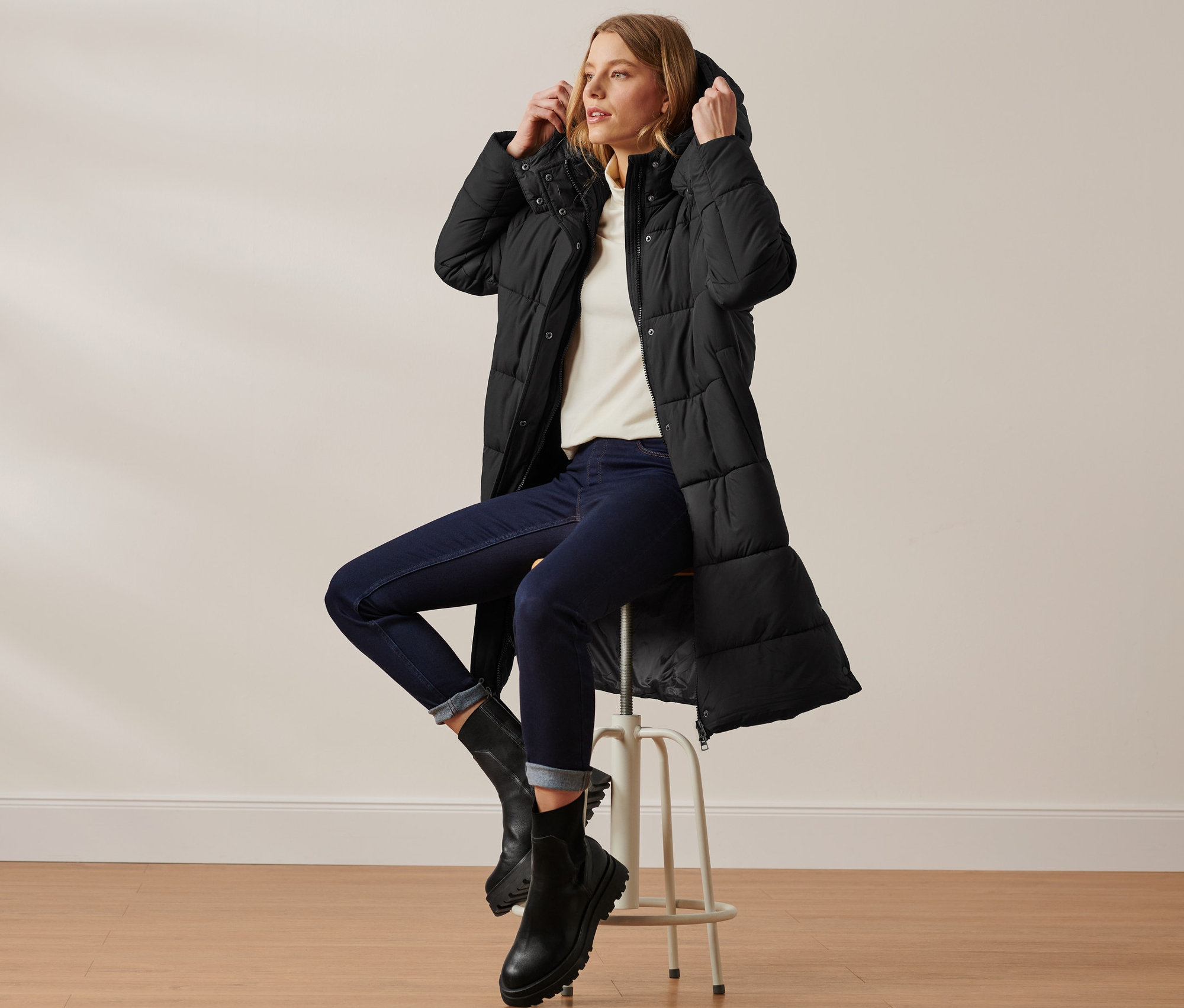 Une femme est assise sur un tabouret et porte un manteau matelassé à capuche noir.