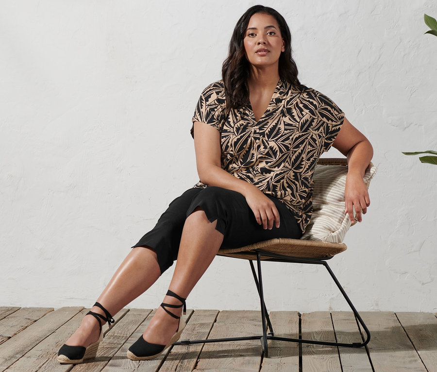 Une femme est assise sur une chaise et porte des espadrilles à talon compensé, un corsaire stretch et un t-shirt imprimé à col montant.