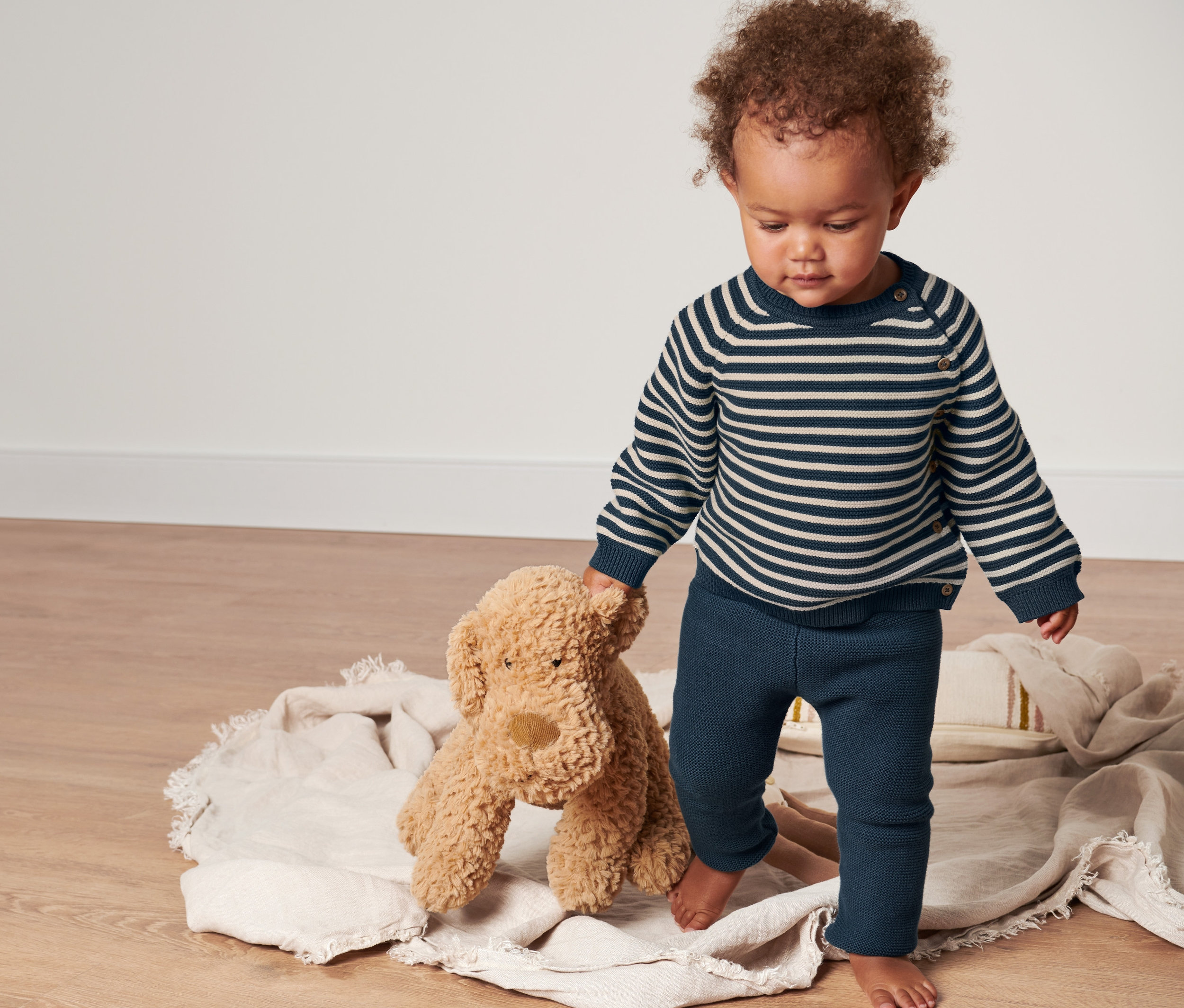 Un bébé se tient debout sur une couverture et tient un ours en peluche, il porte un ensemble en tricot pour bébé.