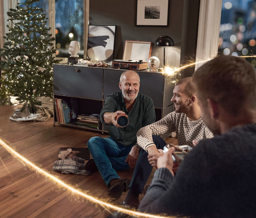 Trois hommes sont assis par terre dans un salon, l'un tenant une Enceinte Bluetooth® vintage avec housse en similicuir. Ils portent un jean, un pull en maille et une Chemise en flanelle.