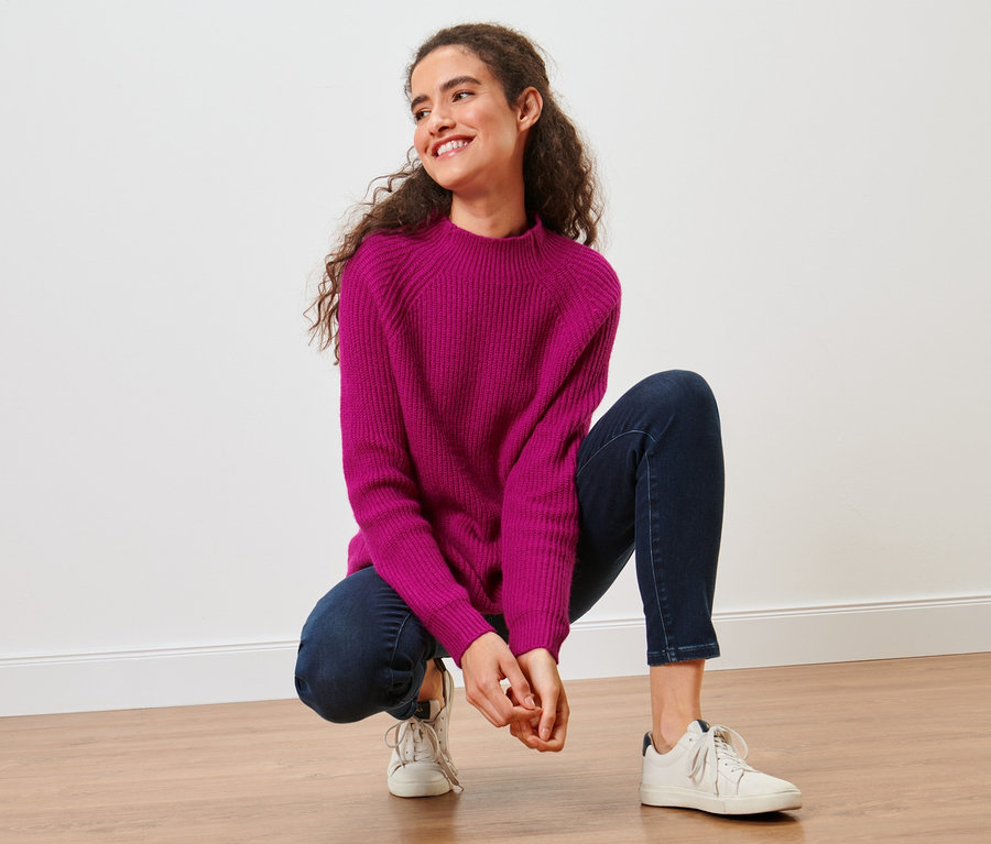 Femme accroupie portant un pull en maille rose à col montant.