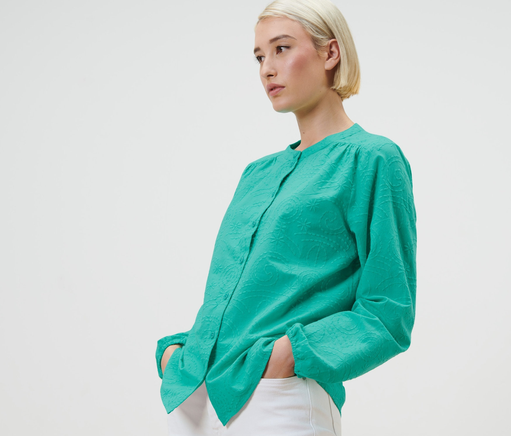 Une femme blonde pose en chemisier à broderie vert avec un pantalon blanc.