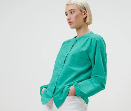 Une femme blonde pose en chemisier à broderie vert avec un pantalon blanc.