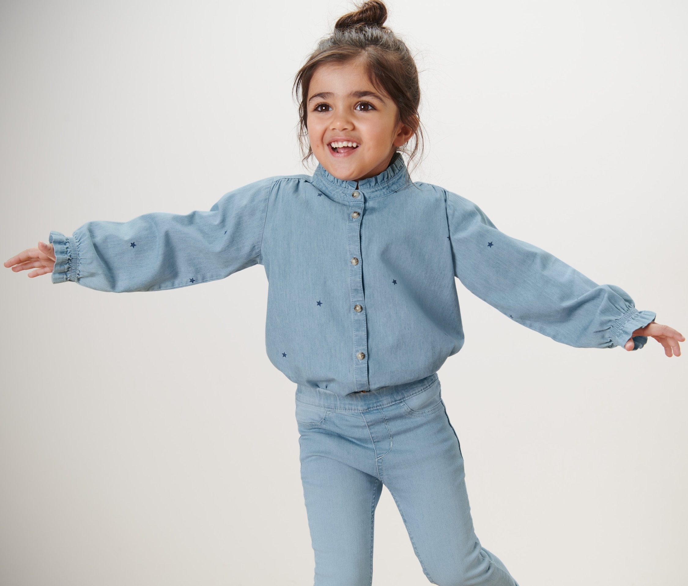Une fillette souriante aux bras écartés porte une chemise en chambray pour enfant et un jean.