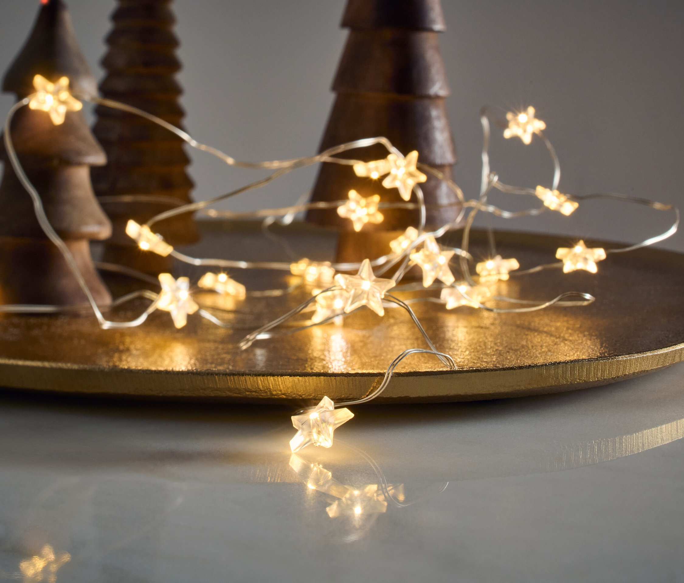 Micro-guirlande lumineuse à LED, étoiles, posée sur un plateau doré à côté de sapins décoratifs en bois.