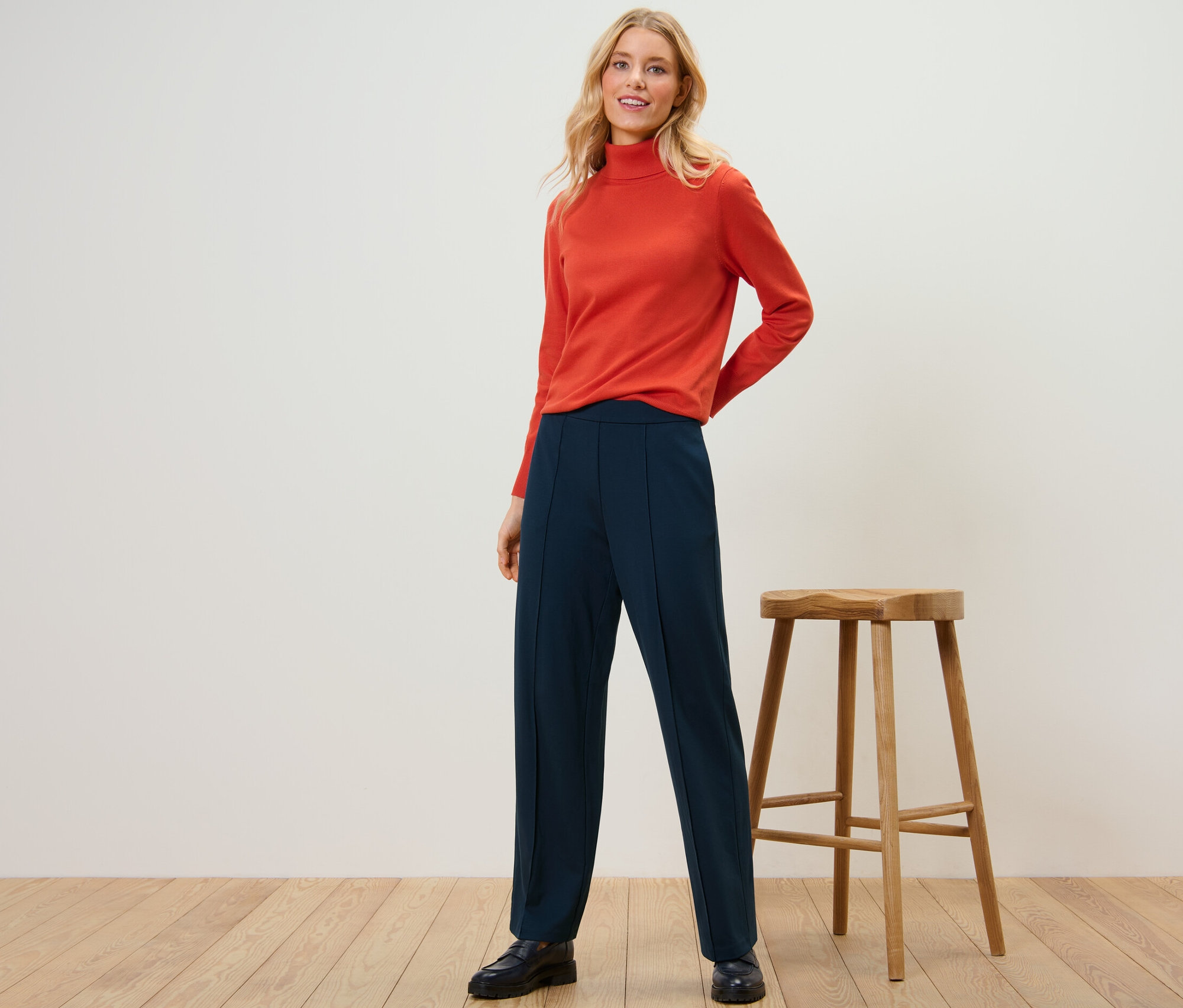Une femme pose devant un fond de studio, portant un pull à col roulé orange et un pantalon en jersey bleu marine avec passepoil.