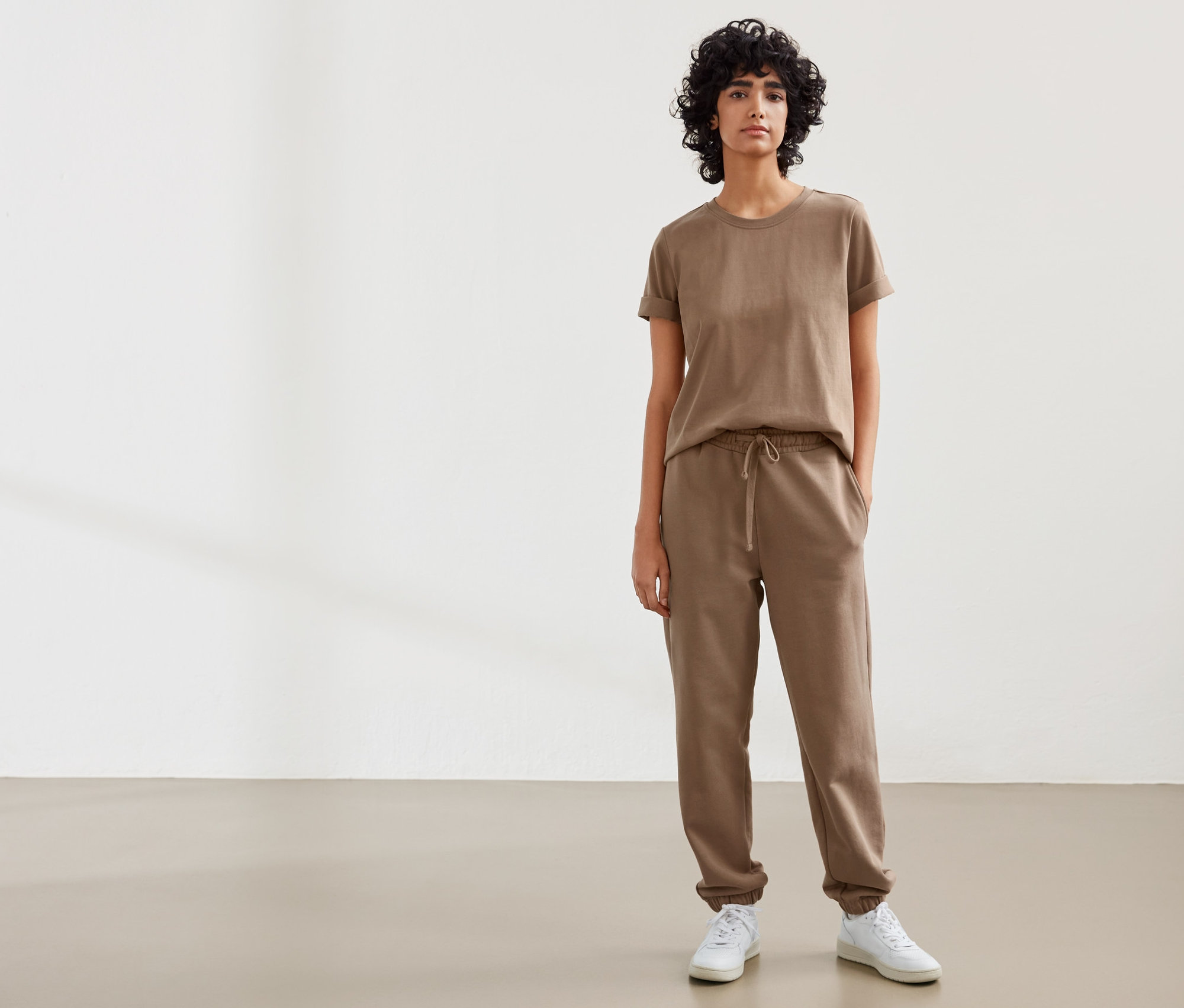Une femme debout porte un t-shirt NAH/STUDIO Heavy Weight Shirt en coton durable couleur noisette et un pantalon de jogging NAH/STUDIO en coton durable couleur noisette.