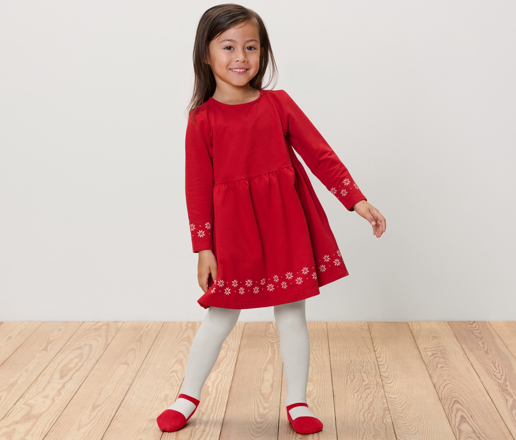 Une fillette souriante pose dans une robe rouge à manches longues avec des flocons de neige et des collants blancs. 2 collants thermiques pour enfant, ballerine.