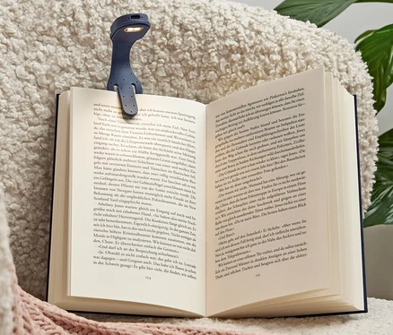 Une mini lampe de lecture LED bleue avec batterie est fixée sur un livre ouvert posé sur un fauteuil.