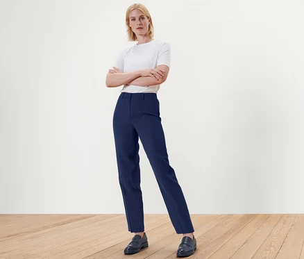 Une femme blonde se tient les bras croisés, portant un pantalon tissé bleu et des chaussures sur un plancher de bois.