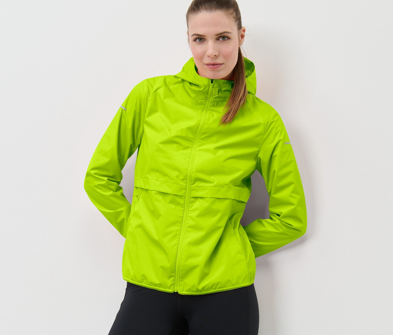 Une femme pose sur un fond blanc et porte une veste de course coupe-vent verte fluo avec des leggings noirs.