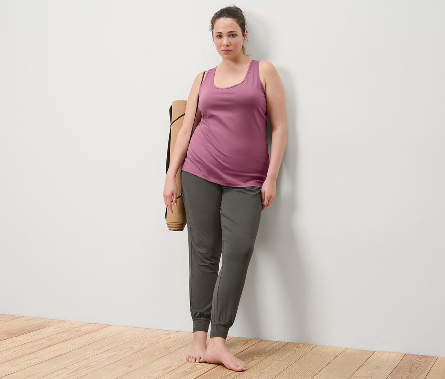 Femme adossée au mur portant 2 tops, mauve, et un pantalon de yoga, un tapis de yoga sur l'épaule.