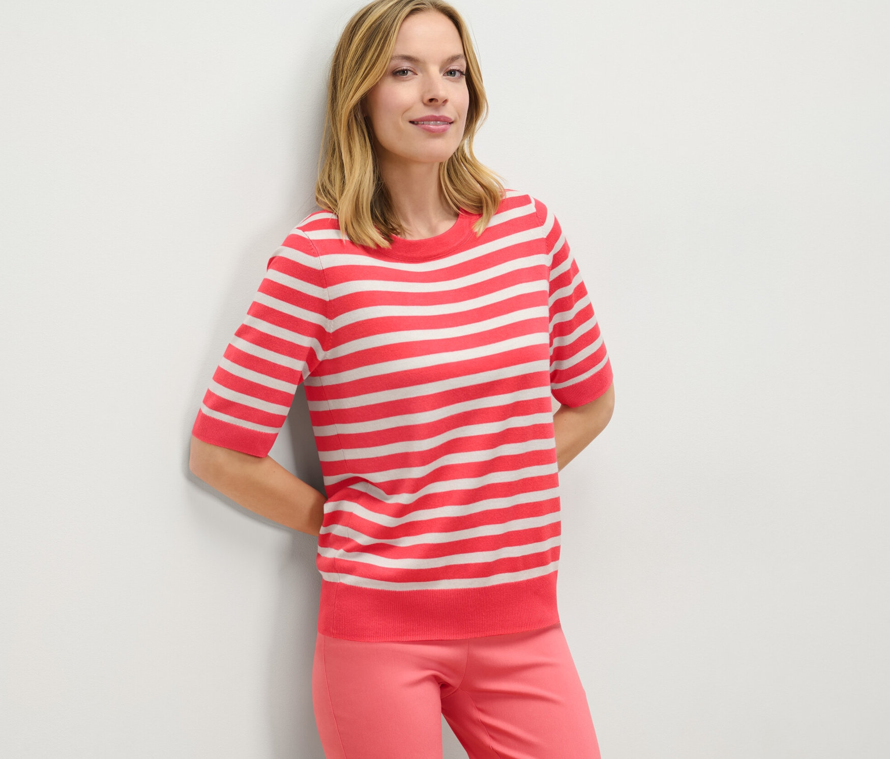 Femme appuyée contre un mur portant un pull en maille fine rayé et un pantalon rose.