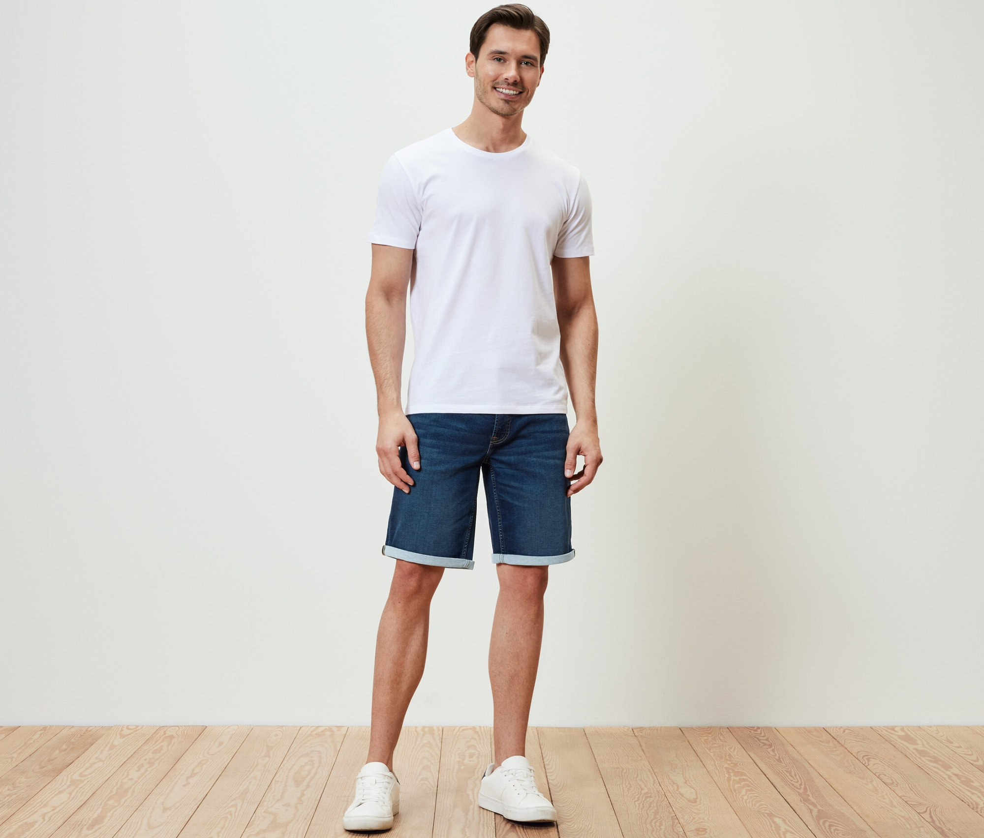 Un homme debout porte un short en jean confortable, un t-shirt blanc et des baskets.