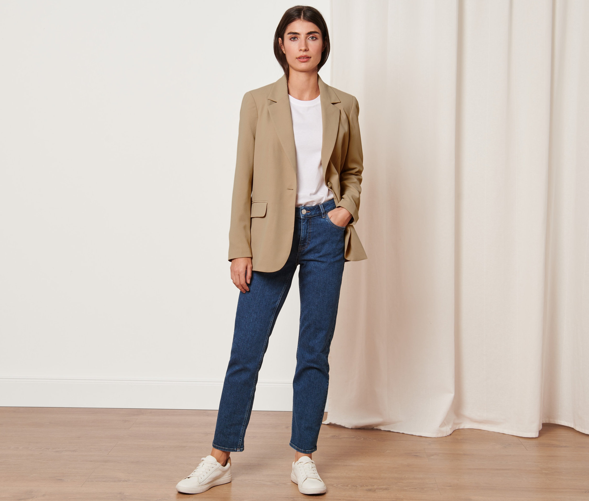 Une femme pose avec un blazer en toile marron clair, un jean bleu, un t-shirt blanc et des baskets blanches.