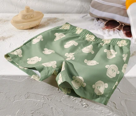 Un boardshort pour enfant vert à motifs de singes est posé sur une surface blanche à côté d'un bateau jouet, d'une serviette, de lunettes de soleil et de crème solaire.