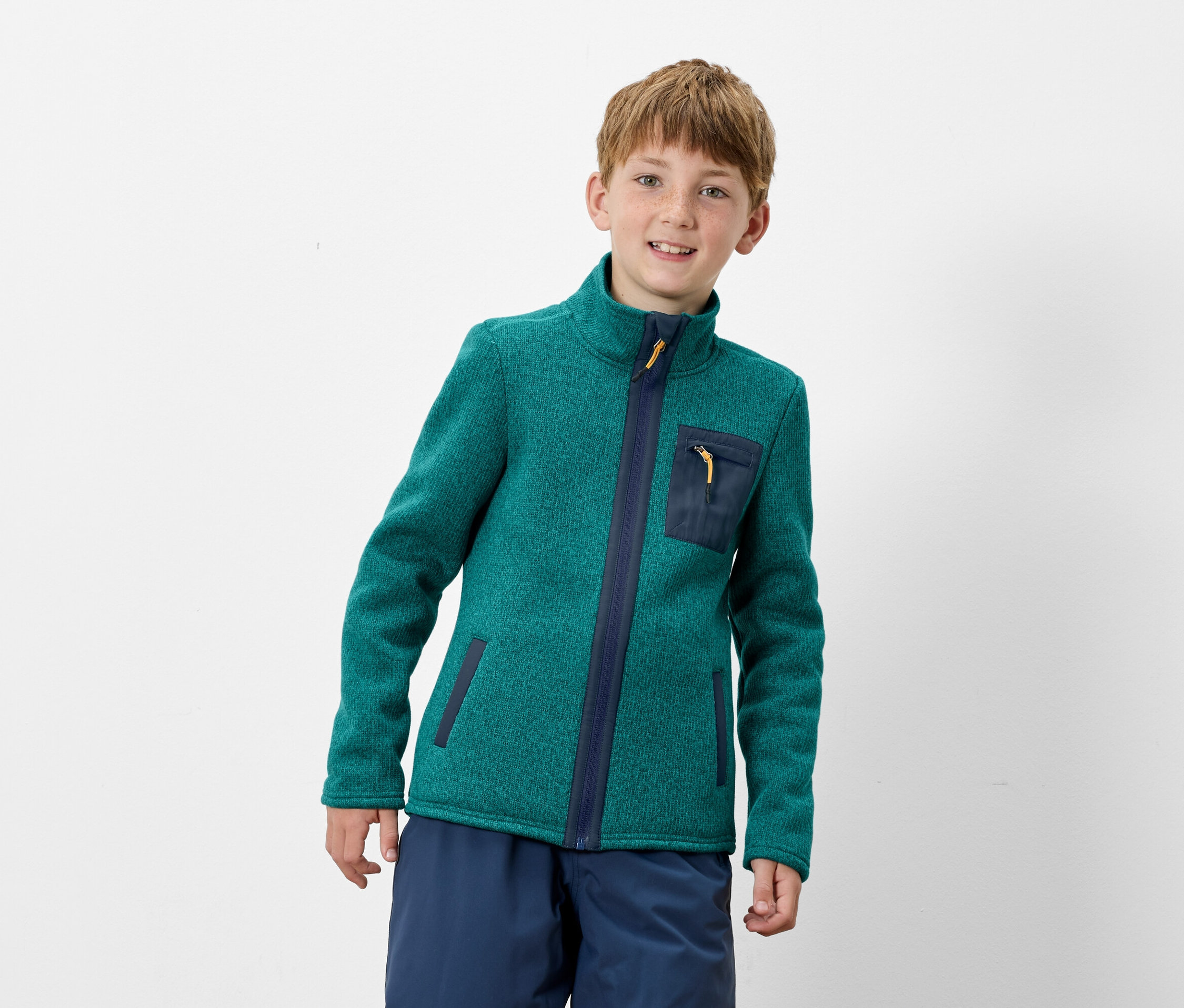 Un garçon porte une veste polaire verte zippée avec des détails bleu foncé et un pantalon bleu.