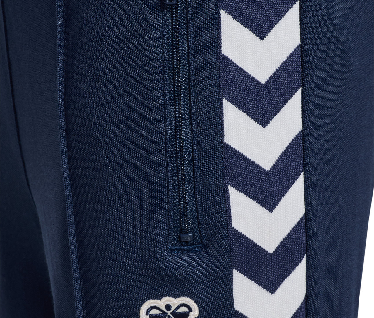 Gros plan sur un pantalon bleu marine HUMMEL HMLJR Archive Regular Poly Pants avec fermeture éclair et logo blanc.