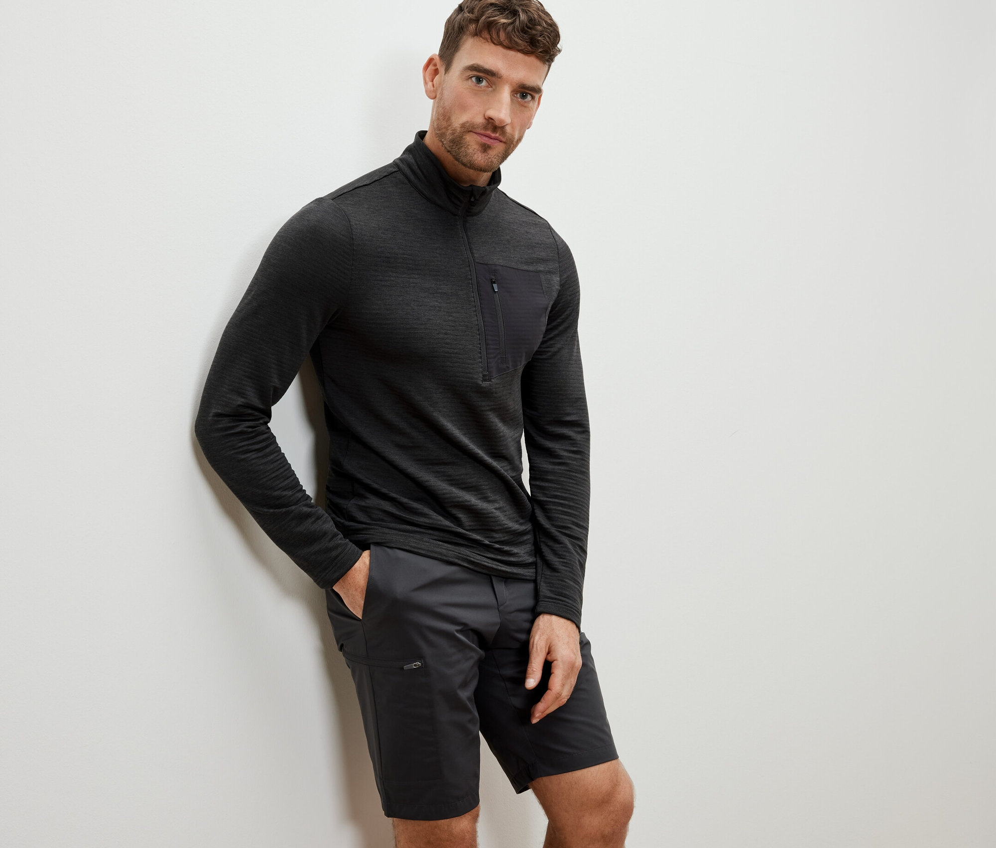 Un homme appuyé contre un mur portant une chemise fonctionnelle outdoor anthracite et un short.
