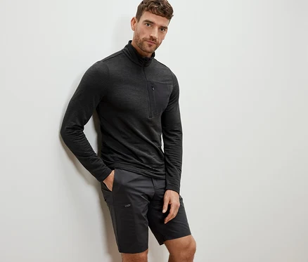 Un homme appuyé contre un mur portant une chemise fonctionnelle outdoor anthracite et un short.