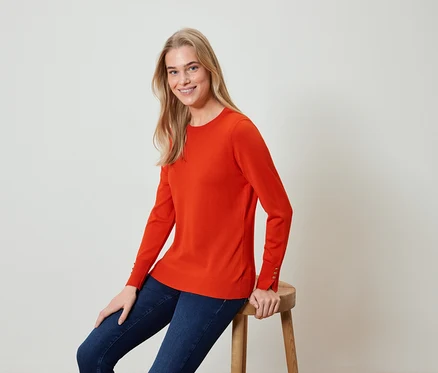 Une femme blonde est assise sur un tabouret et porte un pull orange et un jean bleu.