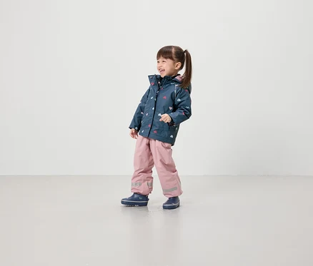 Une fillette souriante portant une veste à motifs de cœurs, un pantalon rose et des chaussures bleues se tient sur un fond clair.