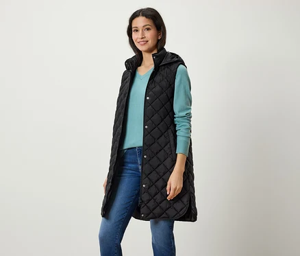 Une femme porte un long gilet noir matelassé à capuche, un pull bleu et un jean.