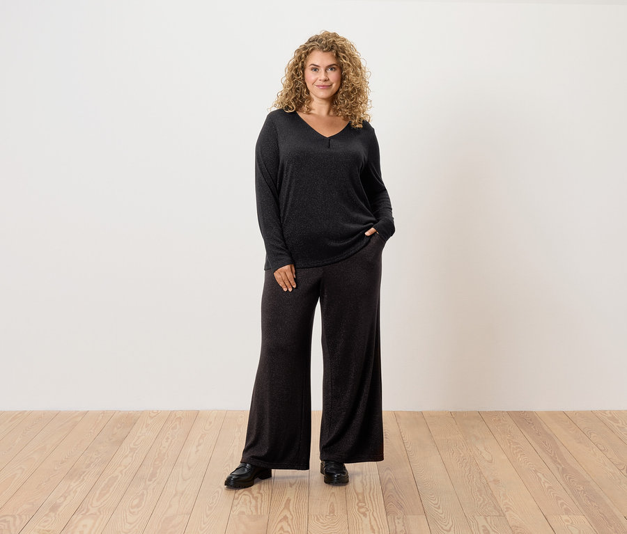 Femme aux cheveux bouclés posant en pull noir et pantalon large.