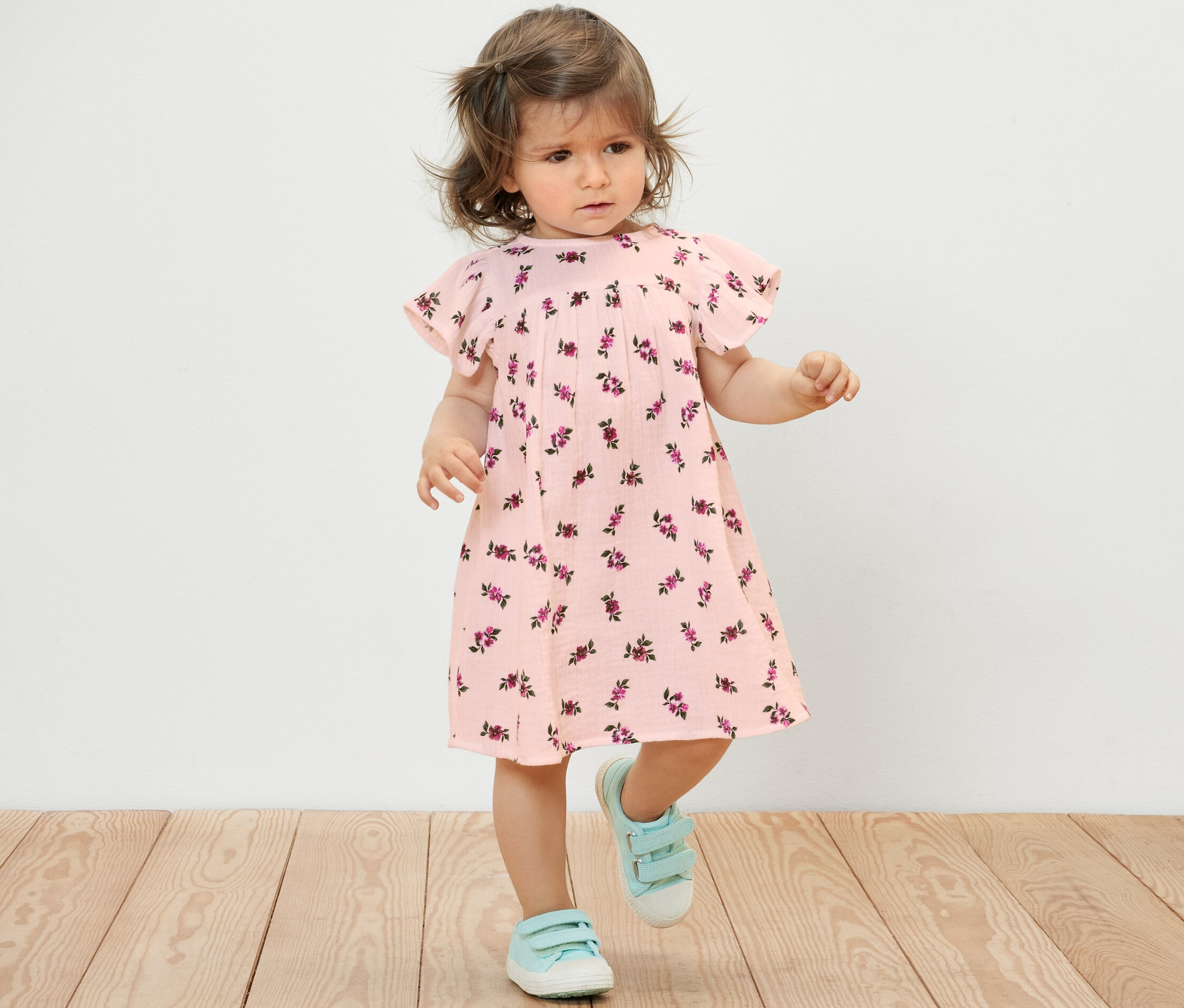 Une petite fille portant une robe en mousseline pour bébé à fleurs et des chaussures bleues se tient sur un plancher en bois.