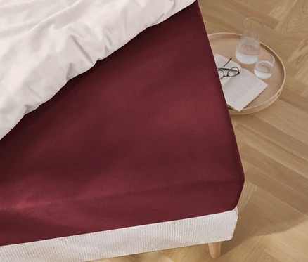 Gros plan d'un drap-housse rouge sur un lit, à côté duquel se trouve une table ronde avec des lunettes, un livre et un verre d'eau.