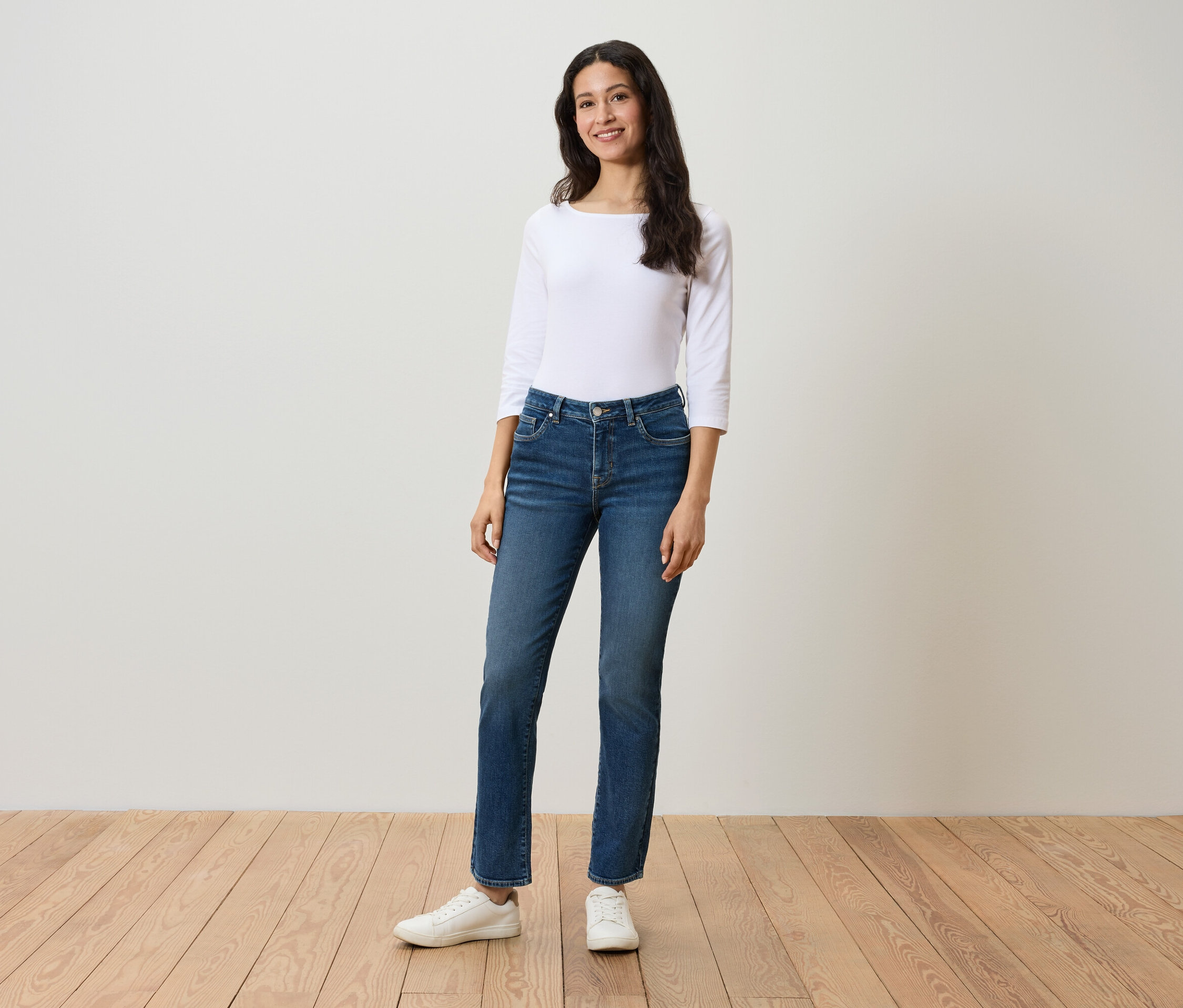 Une femme se tient sur un plancher en bois et porte un chemisier blanc à manches trois-quarts, un jean bleu et des baskets blanches.