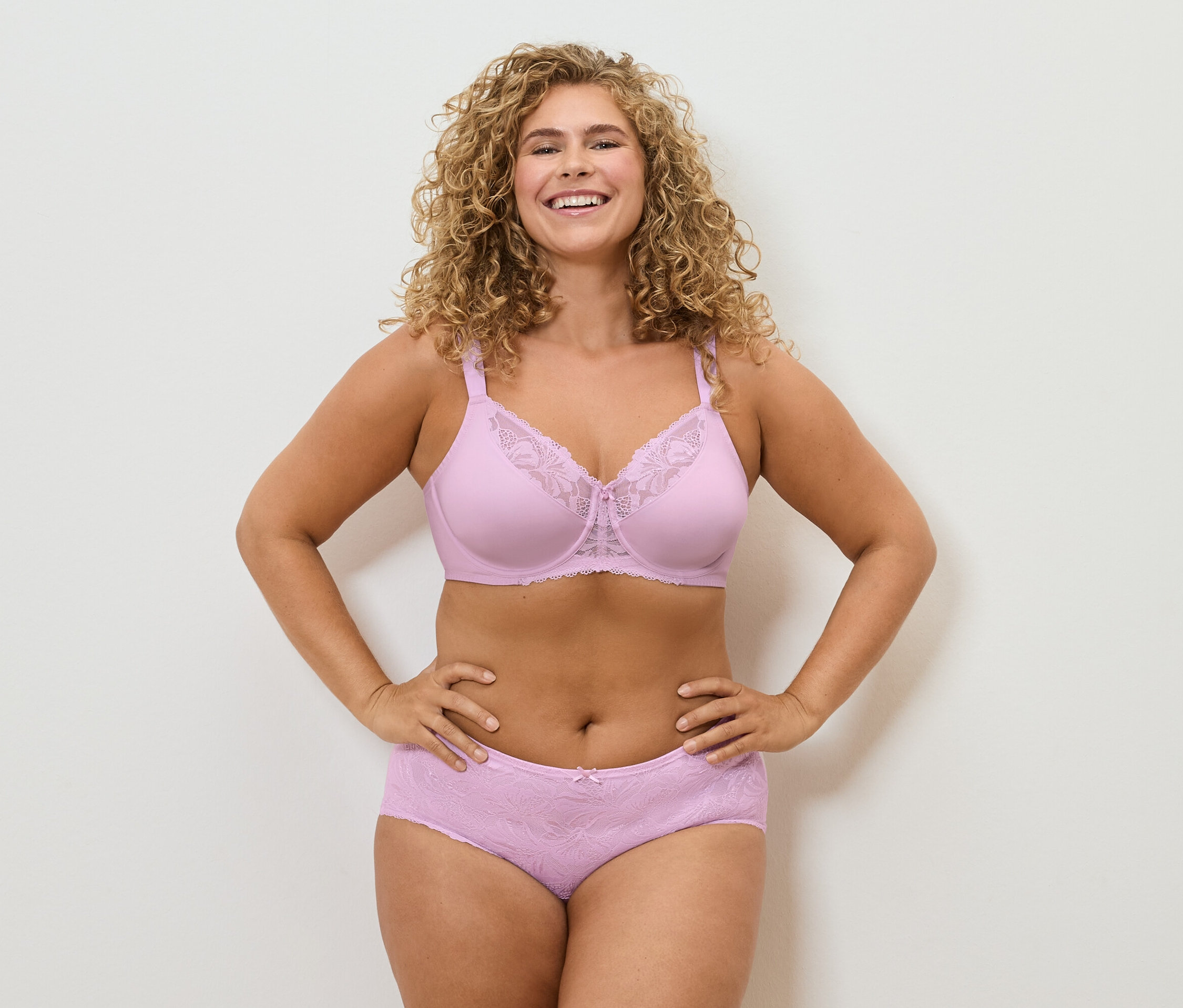 Une femme souriante aux cheveux bouclés pose en soutien-gorge et culotte en dentelle rose.