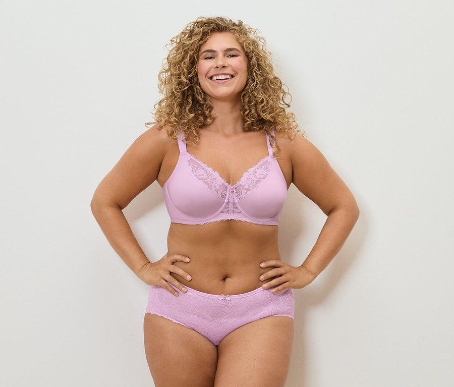 Une femme souriante aux cheveux bouclés pose en soutien-gorge et culotte en dentelle rose.