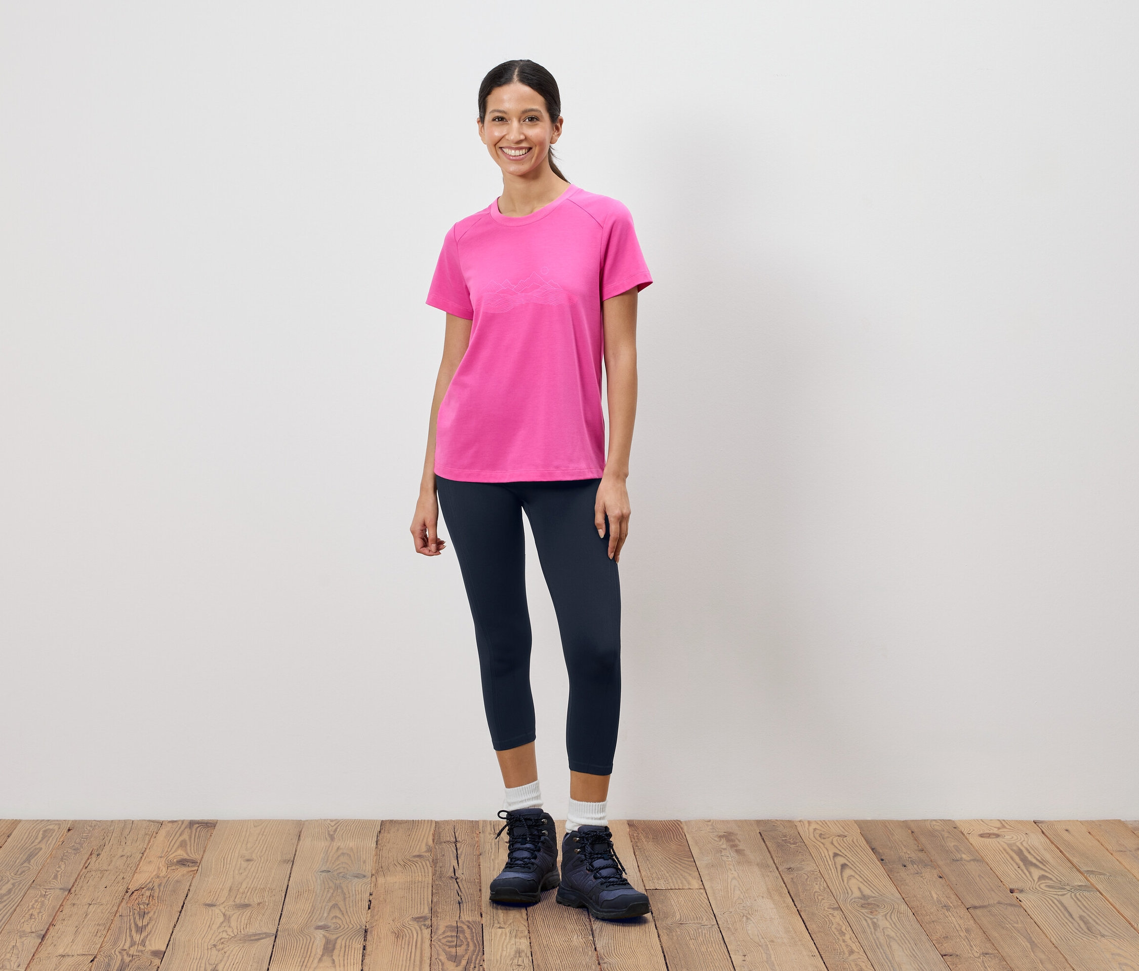 Une femme se tient devant un mur blanc et porte un t-shirt rose, un legging foncé et des chaussures de randonnée.