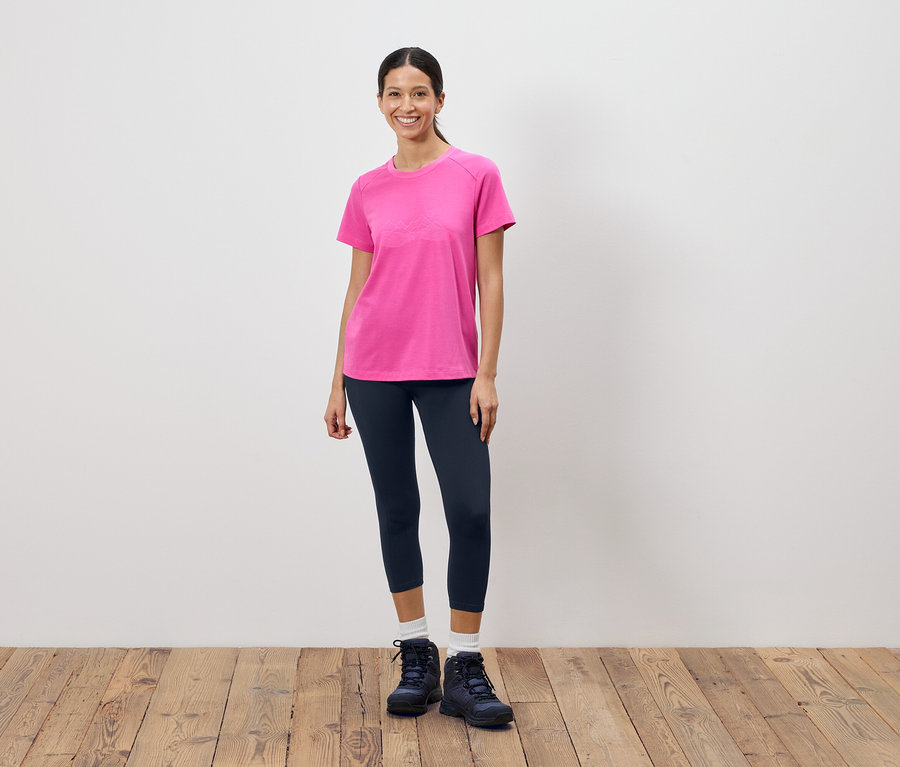 Une femme se tient devant un mur blanc et porte un t-shirt rose, un legging foncé et des chaussures de randonnée.