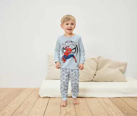 Un garçon en pyjama Spiderman se tient sur un plancher en bois devant un canapé blanc.