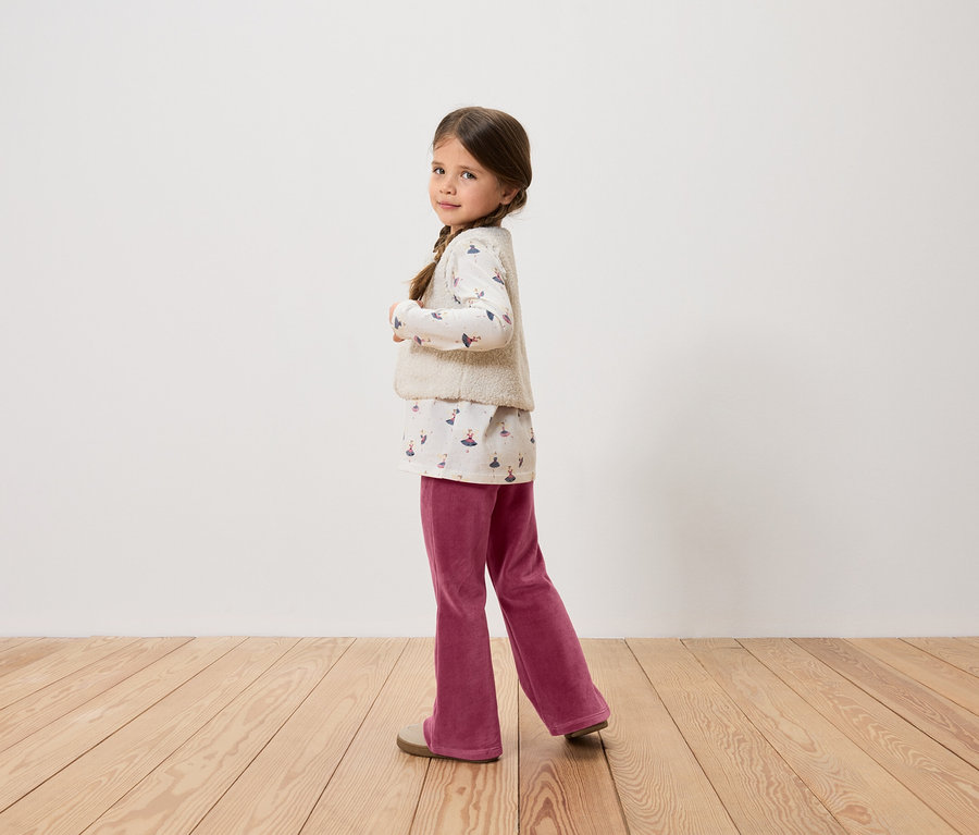 Une fillette aux cheveux bruns tressés pose en haut imprimé, gilet, pantalon bordeaux et chaussures.
