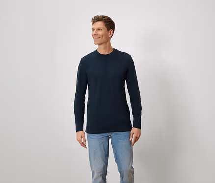 Un homme aux cheveux bruns et aux yeux bleus porte un t-shirt bleu à manches longues et un jean.