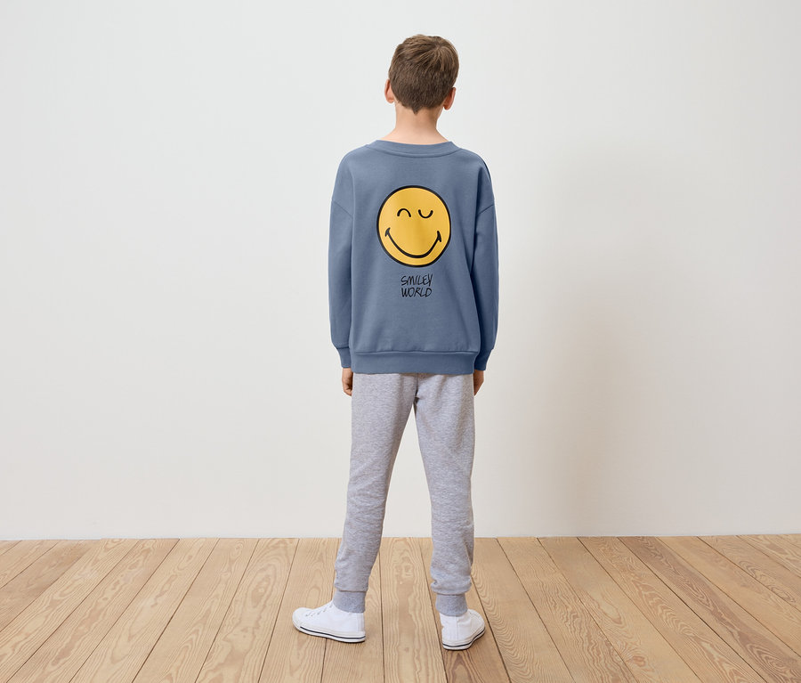 Un garçon, vu de dos, porte un sweat-shirt bleu avec un smiley jaune et un pantalon de survêtement gris.