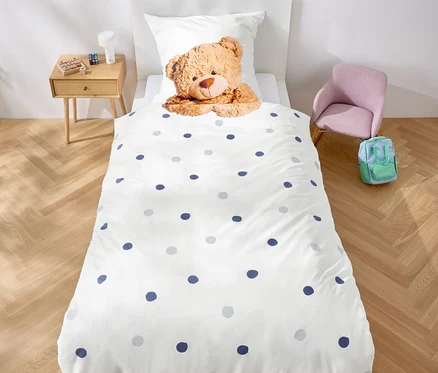 Dans la chambre, il y a un lit avec des draps blancs à pois bleus et un ours en peluche. Une guirlande de triangles violets est accrochée au-dessus du lit.