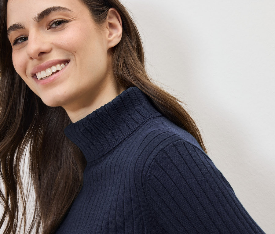 Une femme souriante aux longs cheveux bruns porte un pull à col roulé bleu marine.