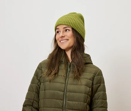 Une femme souriante porte une veste matelassée olive et un bonnet tricoté.