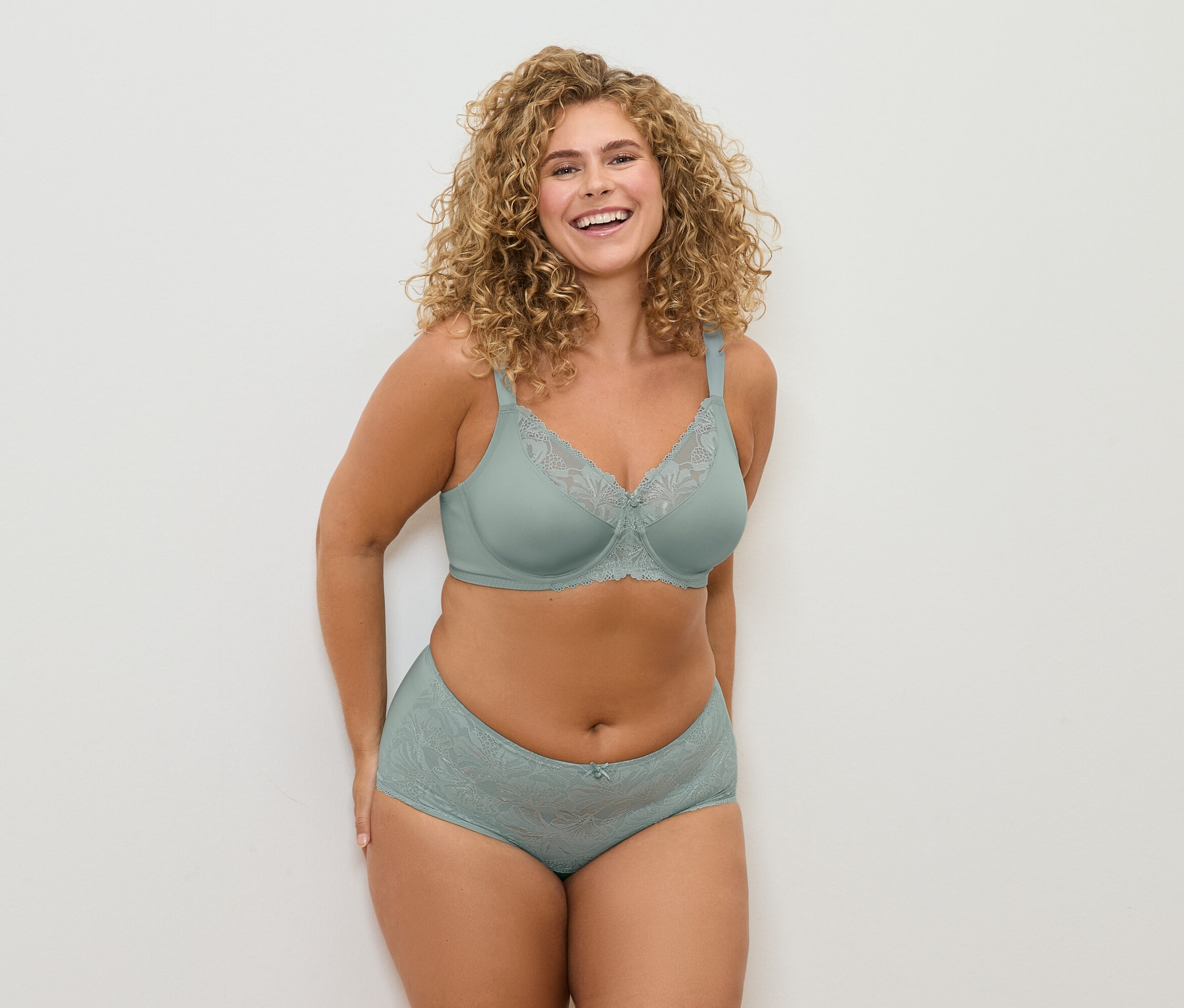 Femme souriante aux cheveux bouclés posant dans un soutien-gorge vert d'eau avec de la dentelle et une culotte assortie.