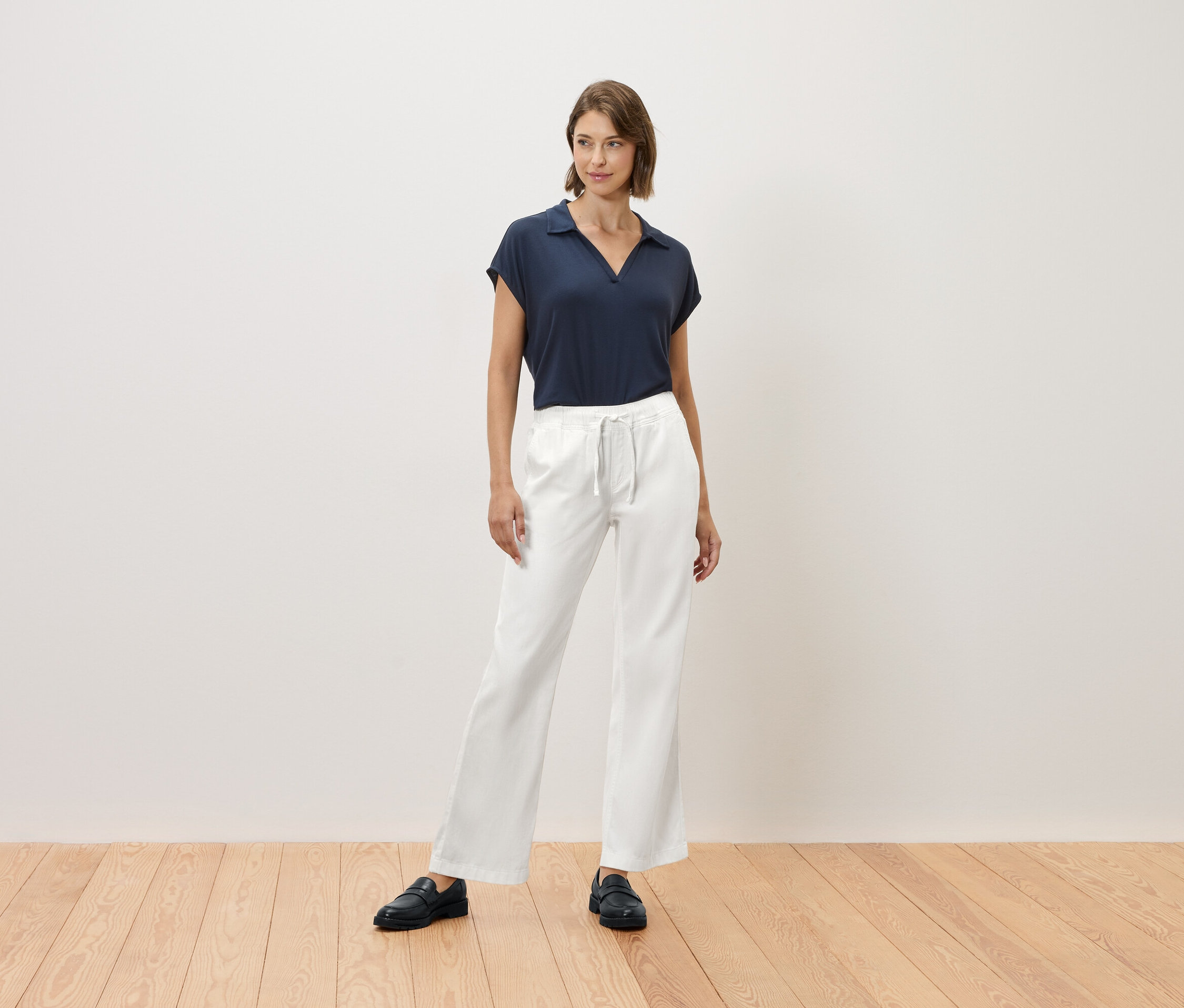 Une femme se tient sur un plancher en bois et porte un t-shirt bleu marine et un pantalon blanc.
