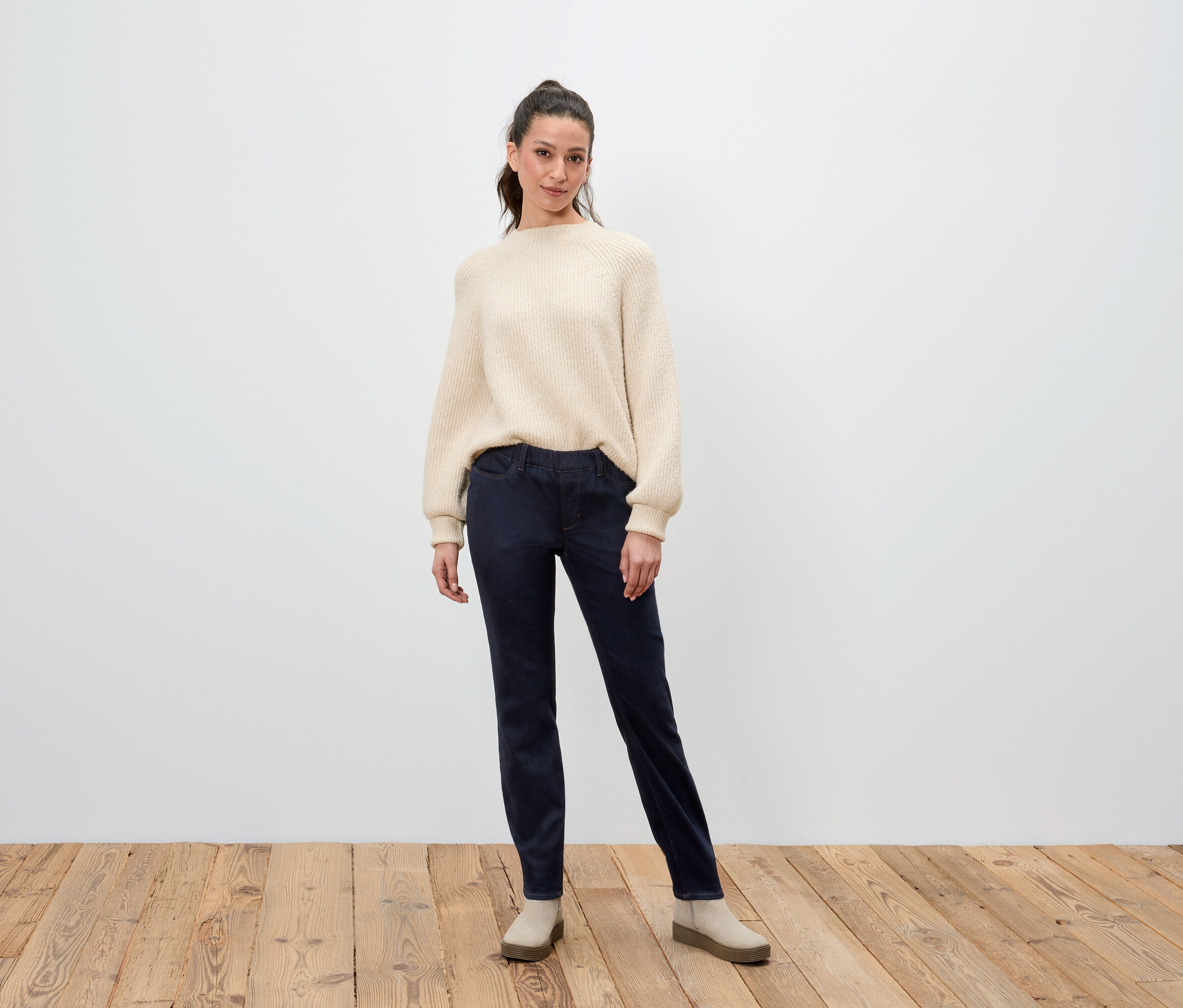 Une femme debout pose en pull crème, pantalon bleu marine et chaussures beiges.