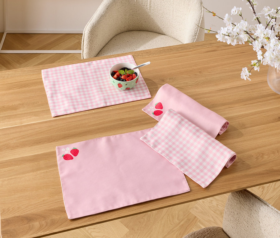 Sur une table en bois se trouvent des sets de table roses, un avec un motif de fraises, un bol de baies et un vase avec des branches fleuries.